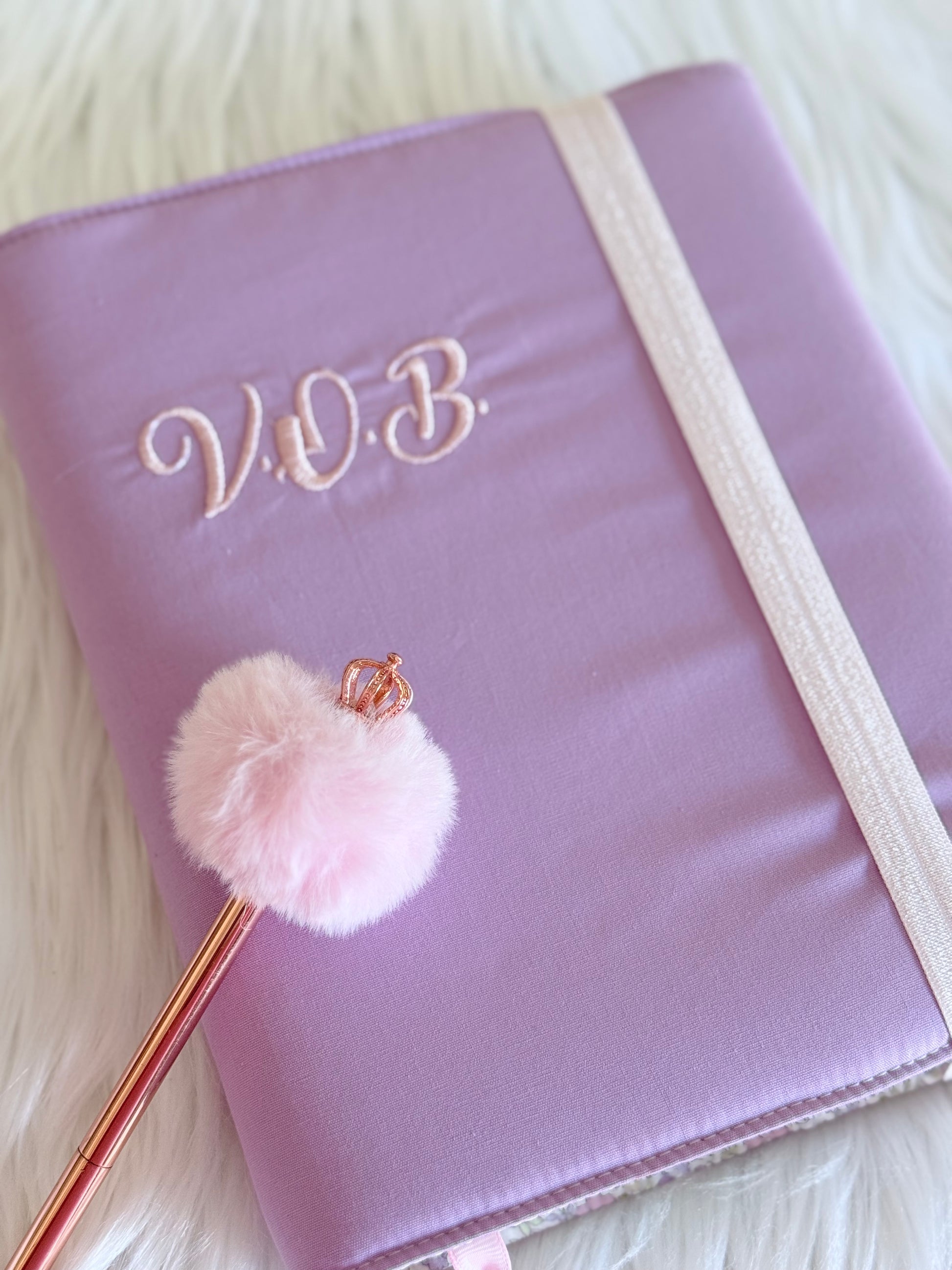 Funda de tela ajustable lila con bordado personalizado de iniciales y marcapáginas de pompón por Salud Ruiz Hecho a Mano.⁠