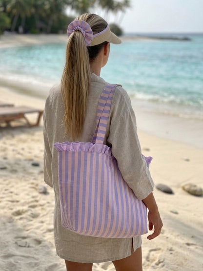 "Bolso de playa de tela con volantes de rayas lavanda colgado al hombro de una mujer en la playa"